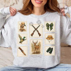 Peut inclure: Un sweat-shirt gris clair avec des illustrations sur le th&egrave;me de la chasse. Les motifs comprennent un chien avec un canard, des fusils crois&eacute;s, un n&oelig;ud vert, un chapeau, un canard, un couteau, un cerf et des jumelles. Le texte "Brushed Cafe" est imprim&eacute; en haut &agrave; droite.