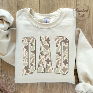 K&ouml;nnte beinhalten: Cremefarbenes Sweatshirt mit dem Wort "DAD" in gro&szlig;en Blockbuchstaben. Die Buchstaben sind mit einem Camouflagemuster in Braun-, Gr&uuml;n- und Beiget&ouml;nen gef&uuml;llt. Das Sweatshirt hat einen Rundhalsausschnitt und lange &Auml;rmel. Der Text "Brushed Cafe" ist sichtbar.