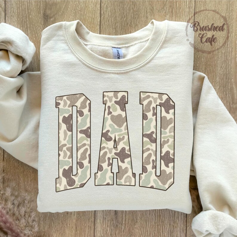 K&ouml;nnte beinhalten: Cremefarbenes Sweatshirt mit dem Wort "DAD" in gro&szlig;en Blockbuchstaben. Die Buchstaben sind mit einem Camouflagemuster in Braun-, Gr&uuml;n- und Beiget&ouml;nen gef&uuml;llt. Das Sweatshirt hat einen Rundhalsausschnitt und lange &Auml;rmel. Der Text "Brushed Cafe" ist sichtbar.