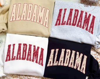 May include: Four crewneck sweatshirts in beige, light gray, white, and black. Each sweatshirt features the word ALABAMA printed in a distressed, arched collegiate font across the chest. The text is dark red on the beige and gray shirts.