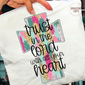 May include: White t-shirt featuring a colorful cross design with the text "Trust in the Lord with all your heart." The cross has pink, teal, and gold brushstroke patterns, with black and white polka dot accents.