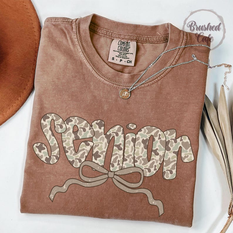 Pode incluir: Uma camiseta Comfort Colors marrom com a palavra "SENIOR" em um padr&atilde;o de camuflagem e um la&ccedil;o gr&aacute;fico. Um colar de prata com um pingente est&aacute; drapeado sobre a camisa. Um chap&eacute;u marrom &eacute; vis&iacute;vel no canto superior esquerdo.