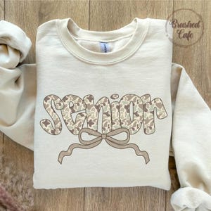 Pode incluir: Camisola creme com a palavra "SENIOR" num padr&atilde;o de camuflagem. Uma fita castanha clara est&aacute; abaixo da palavra. A camisola tem gola redonda e mangas compridas. O texto "Brushed Cafe" est&aacute; no canto superior direito.