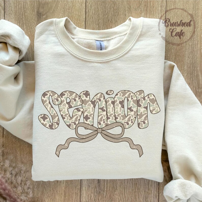 Pode incluir: Camisola creme com a palavra "SENIOR" num padr&atilde;o de camuflagem. Uma fita castanha clara est&aacute; abaixo da palavra. A camisola tem gola redonda e mangas compridas. O texto "Brushed Cafe" est&aacute; no canto superior direito.