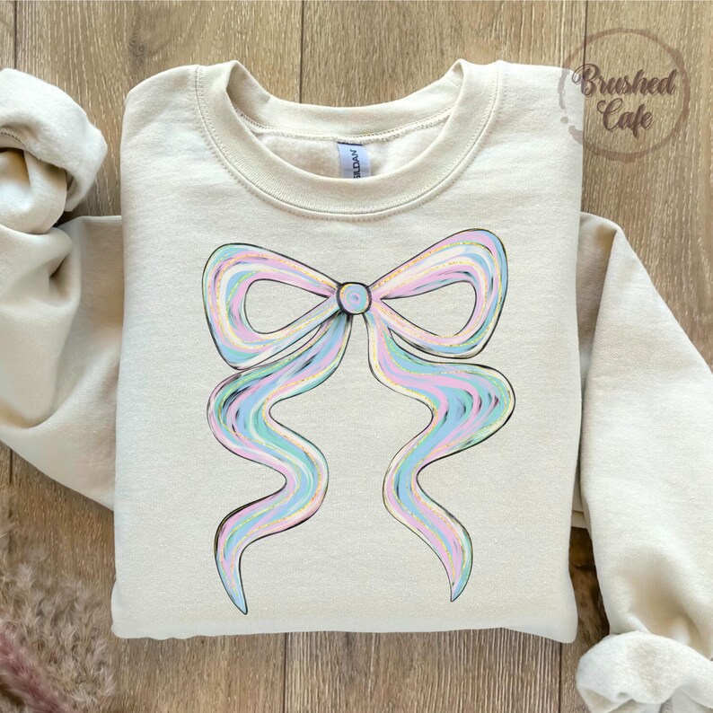 Puede incluir: Sudadera color crema con un dise&ntilde;o de lazo grande en colores pastel. El lazo tiene un efecto acuarela con tonos de azul, rosa y amarillo. La sudadera tiene cuello redondo y mangas largas. El texto "Brushed Cafe" est&aacute; en la esquina superior derecha.