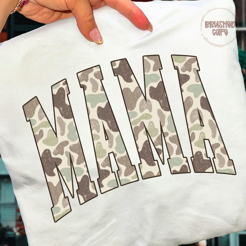 Może przedstawiać: Biała bluza z napisem "MAMA" dużymi, blokowymi literami. Litery mają wz&oacute;r kamuflażu w odcieniach brązu, zieleni i beżu. Bluza jest trzymana przez osobę.