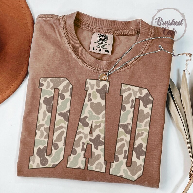 K&ouml;nnte beinhalten: Ein braunes T-Shirt mit dem Wort "DAD" in gro&szlig;en Buchstaben mit einem Camouflage-Muster. Das Shirt hat einen Rundhalsausschnitt und kurze &Auml;rmel. Eine silberne Halskette mit einem kleinen Anh&auml;nger ist &uuml;ber dem Shirt drapiert.