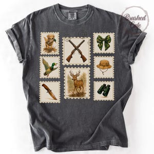 Peut inclure: T-shirt gris fonc&eacute; avec des motifs de timbres-poste sur le th&egrave;me de la chasse. Les timbres repr&eacute;sentent un chien de chasse, des fusils crois&eacute;s, un n&oelig;ud papillon, un chapeau, des jumelles, un couteau, un canard et un cerf. Le t-shirt a un aspect vintage.