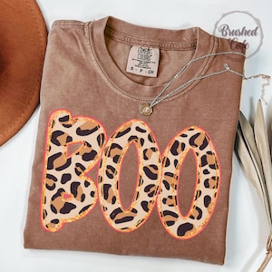 May include: Brown t-shirt with the word "BOO" in a leopard print design with a pink outline. The shirt has a crew neck and short sleeves. A silver necklace with a small pendant is on the shirt.