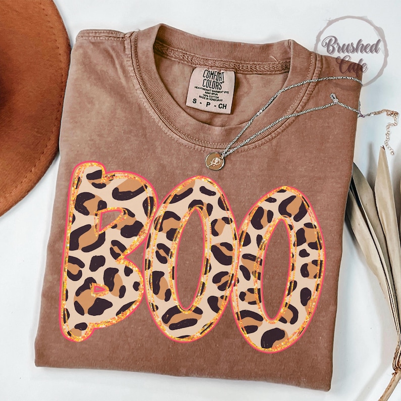 May include: Brown t-shirt with the word "BOO" in a leopard print design with a pink outline. The shirt has a crew neck and short sleeves. A silver necklace with a small pendant is on the shirt.