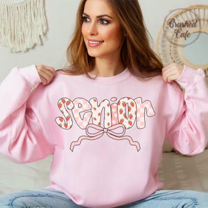 pink sweatshirt with the word 'Senior' in white outlined letters. The letters are decorated with a pink and white checkered pattern and strawberry and flower graphics. A pink bow is below the word.