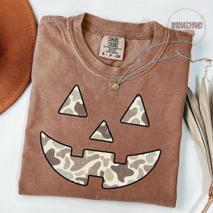 Puede incluir: Una camiseta Comfort Colors marrón con un diseño de cara de calabaza. Los ojos y la boca están delineados en negro y rellenos con un patrón de camuflaje. Un collar plateado con un colgante está sobre la camisa.