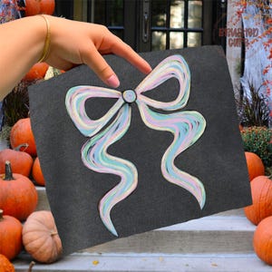 Puede incluir: Un cuadrado gris oscuro con un dise&ntilde;o de lazo en colores pastel. El lazo presenta l&iacute;neas onduladas azules, rosas y amarillas con detalles de purpurina dorada. La imagen est&aacute; sostenida por una mano, con calabazas en el fondo.