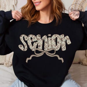 Pode incluir: Camisola preta com a palavra "SENIOR" num padr&atilde;o de camuflagem, com um la&ccedil;o por baixo. A camisola &eacute; de gola redonda.
