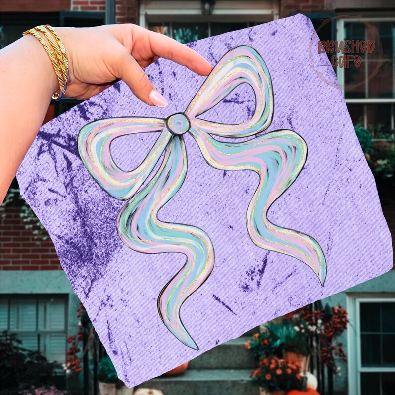 Puede incluir: Un cuadrado lavanda con un dise&ntilde;o de lazo grande en colores pastel. El lazo presenta rayas azules, rosas y amarillas con detalles de purpurina dorada. El fondo tiene un aspecto texturizado y moteado. La imagen est&aacute; sostenida por una mano con pulseras doradas.