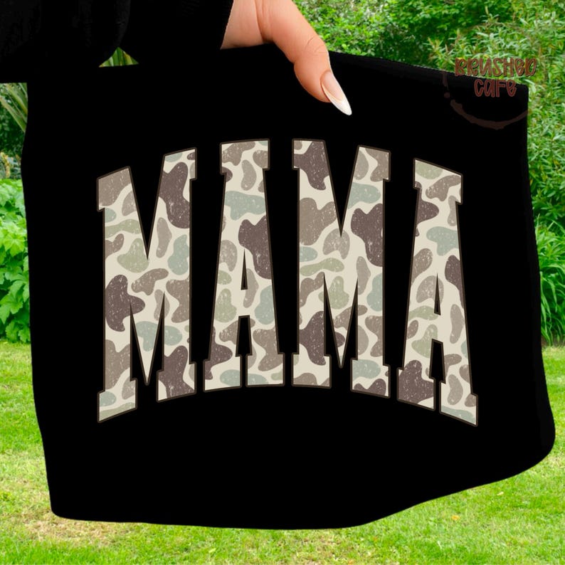 Może przedstawiać: Czarna torba typu tote z napisem "MAMA" w dużych, blokowych literach. Litery wypełnione są wzorem kamuflażu w odcieniach brązu, zieleni i beżu. Torba trzymana jest przez dłoń.