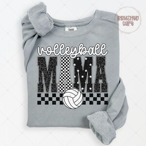May include: A gray sweatshirt with the words "Volleyball Mama" in a mix of fonts and patterns. The word "Volleyball" is in a script font, and "MAMA" is in a bold font with leopard print, checkered, and star patterns. A white volleyball is at the bottom.