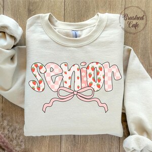 sweatshirt with the word 'Senior' printed on the front in pink and white. The word is decorated with a pink bow and a pattern of pink and white checkered squares, strawberries, and white flowers.