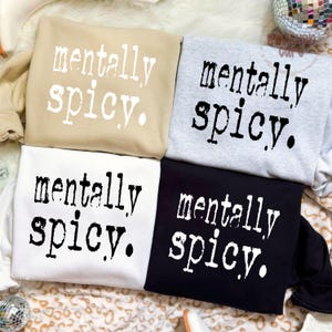 Puede incluir: Cuatro sudaderas en beige, gris, blanco y negro, cada una con la frase "mentally spicy" en una fuente blanca desgastada. Las sudaderas est&aacute;n dobladas y dispuestas juntas, mostrando el dise&ntilde;o.