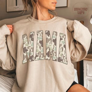 May include: Beige sweatshirt with the word "MAMA" in large, block letters across the front. The letters have a camouflage pattern in shades of green, brown, and gray. The sweatshirt has long sleeves and a crew neck.