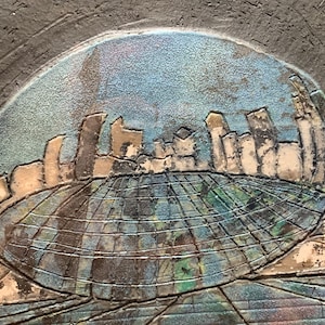 May include: A ceramic wall sculpture with a textured surface. The sculpture depicts a cityscape with buildings and a grid pattern. The background is a blue and green color.