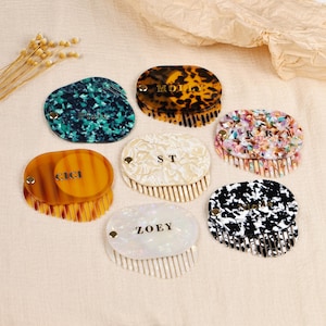 Puede incluir: Una colección de peines ovalados para el cabello en varios colores y patrones, incluyendo carey, verde azulado moteado y blanco y negro. Cada peine presenta una tapa con bisagras con un nombre o iniciales en letras doradas. Los peines están dispuestos sobre una superficie de tono neutro.