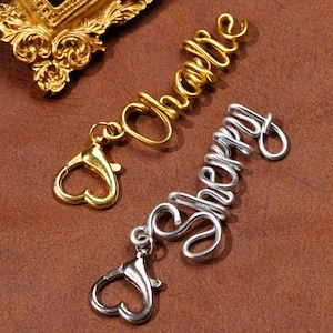 Puede incluir: Dos colgantes de nombre personalizados, uno dorado y otro plateado, cada uno con un cierre de mosquetón. El colgante dorado deletrea "Charlie", y el colgante plateado deletrea "Sherry". Los colgantes son de alambre y están sobre una superficie marrón.