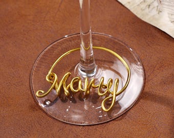May include: A clear wine glass with a gold wire name tag that reads "Marty". The tag is looped around the base of the glass. The background is a brown surface.