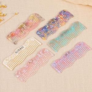 Könnte beinhalten: Sechs farbenfrohe, gemusterte Kämme mit gebogenen Griffen sind auf einer neutralen Oberfläche angeordnet. Jeder Kamm hat ein anderes Farbschema, darunter Rosa, Blau und Gelb. Die Namen "WREN", "KIRAN", "AJ", "JORDAN" und "WE" sind auf den Kämmen aufgedruckt.