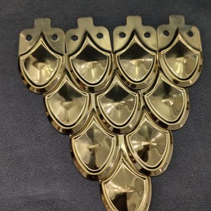 Puede incluir: Una colección de piezas metálicas doradas en forma de escudo, dispuestas en un patrón triangular. Cada pieza tiene una base redondeada y una pequeña pestaña con un agujero en la parte superior. Las piezas tienen una superficie brillante y reflectante.