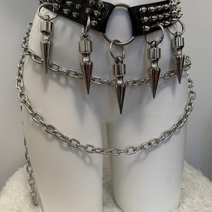 May include: Black leather belt with silver metal spikes and chain accents. The belt features a large metal ring and dangling cone-shaped pendants. A silver chain hangs from the belt, adding to the edgy aesthetic. The belt is displayed on a white mannequin.