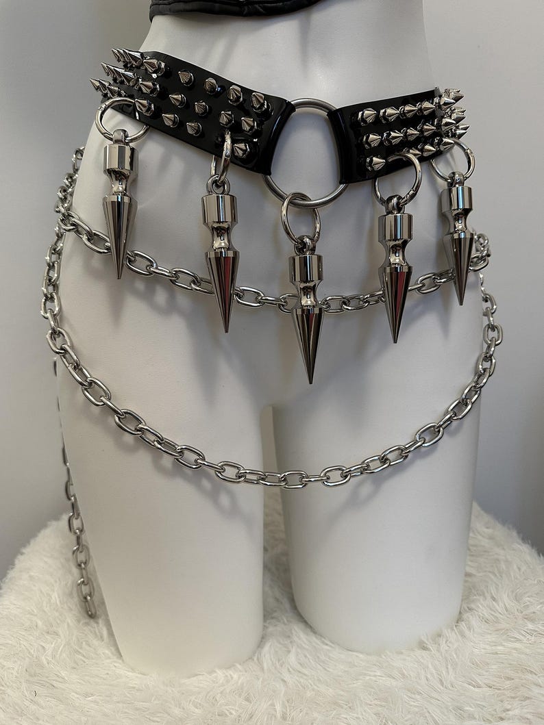May include: Black leather belt with silver metal spikes and chain accents. The belt features a large metal ring and dangling cone-shaped pendants. A silver chain hangs from the belt, adding to the edgy aesthetic. The belt is displayed on a white mannequin.