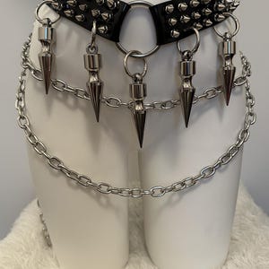May include: A black leather choker with silver spikes and a large silver ring. Dangling from the choker are five silver cone-shaped pendants. A silver chain is attached to the choker and hangs down.