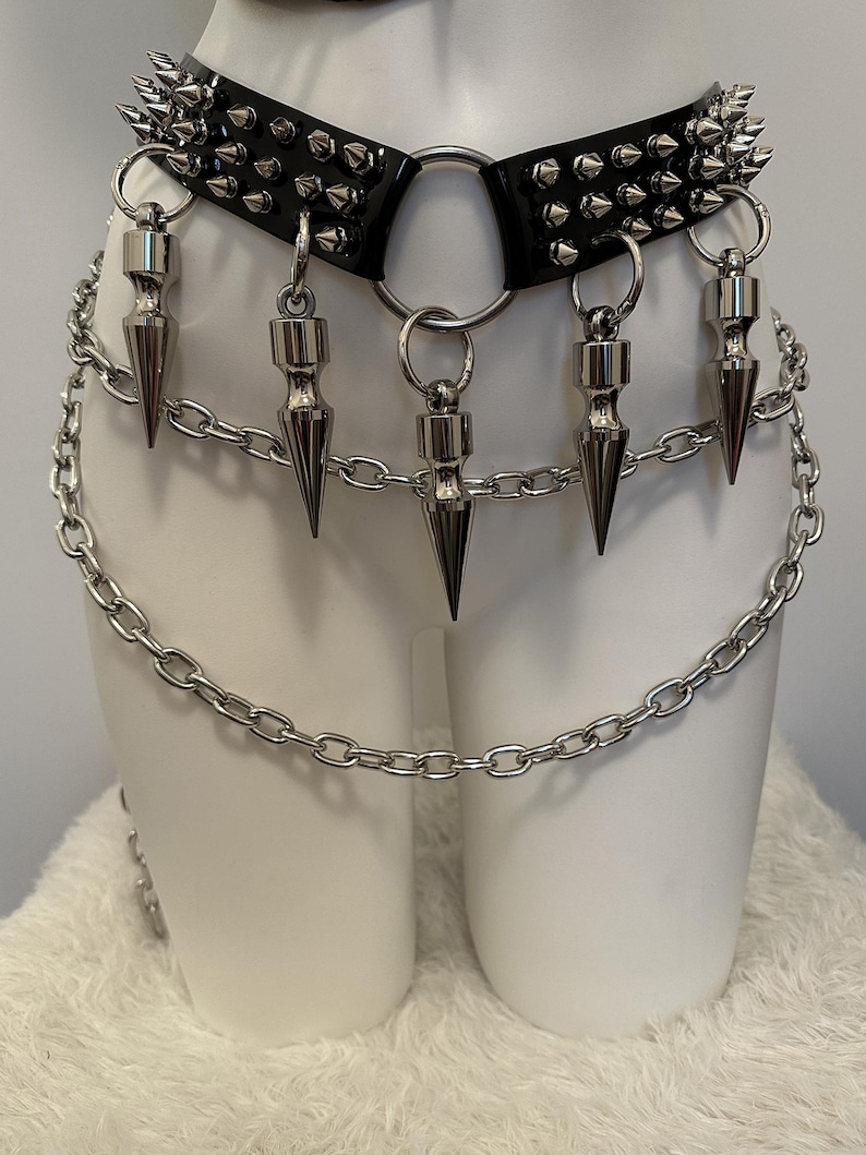 May include: A black leather choker with silver spikes and a large silver ring. Dangling from the choker are five silver cone-shaped pendants. A silver chain is attached to the choker and hangs down.