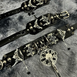 Puede incluir: Tres collares de cuero negro con detalles en metal plateado. Cada collar presenta un dise&ntilde;o de cruz, pinchos y cuentas. El collar m&aacute;s grande tiene un gran colgante de cruz plateada con una figura de Jes&uacute;s.