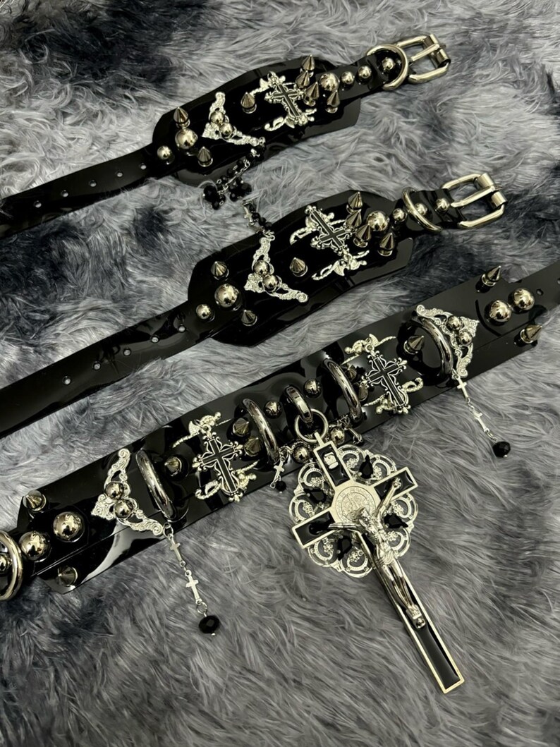Puede incluir: Tres collares de cuero negro con detalles en metal plateado. Cada collar presenta un dise&ntilde;o de cruz, pinchos y cuentas. El collar m&aacute;s grande tiene un gran colgante de cruz plateada con una figura de Jes&uacute;s.