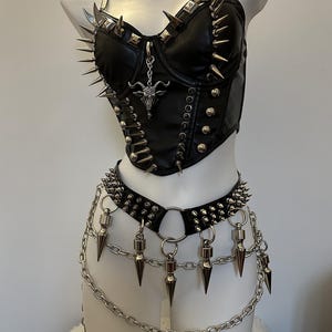 May include: Black faux leather corset and belt set with silver spikes and chain details. The corset features a pendant with a goat head. The belt has dangling spikes and a chain that hangs down.