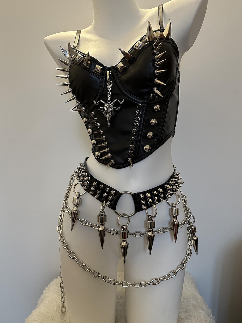 May include: Black faux leather corset and belt set with silver spikes and chain details. The corset features a pendant with a goat head. The belt has dangling spikes and a chain that hangs down.