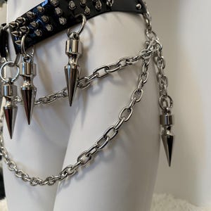 May include: Black leather belt with silver spikes and chain accents. The belt features multiple rows of silver spikes and dangling silver cone-shaped charms. A silver chain hangs from the belt, adding a punk rock aesthetic.