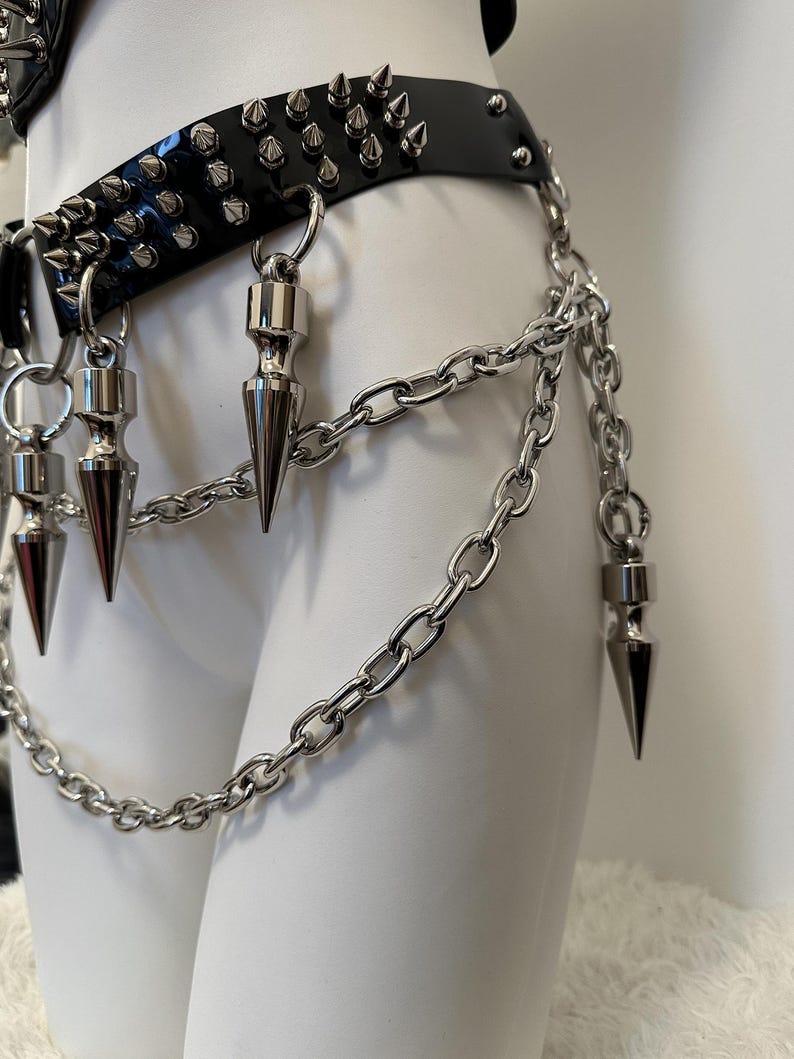 May include: Black leather belt with silver spikes and chain accents. The belt features multiple rows of silver spikes and dangling silver cone-shaped charms. A silver chain hangs from the belt, adding a punk rock aesthetic.