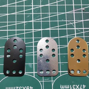 Könnte beinhalten: Drei Metallplatten mit mehreren Löchern. Die Platten sind schwarz, silber und goldfarben. Die schwarze Platte misst 49x32mm, die silberne Platte befindet sich in der Mitte und die goldfarbene Platte misst 47x31mm. Die Platten liegen auf einer grünen Schneidematte.