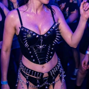 May include: Black leather corset and matching bottoms with silver spikes and chain detailing. The outfit features a decorative pendant and is worn in a crowded, dimly lit environment, suggesting a club or concert setting.
