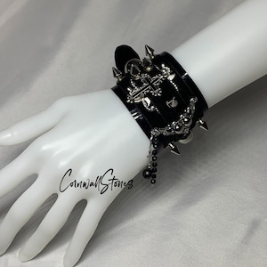 Puede incluir: Un brazalete de cuero negro con tachuelas plateadas, un dije de cruz y una cadena de cuentas. El brazalete se lleva en la mu&ntilde;eca de un maniqu&iacute;.