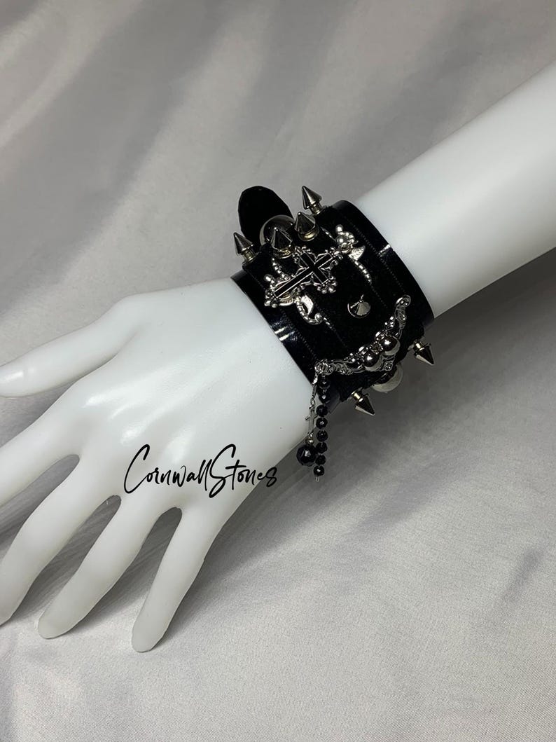 Puede incluir: Un brazalete de cuero negro con tachuelas plateadas, un dije de cruz y una cadena de cuentas. El brazalete se lleva en la mu&ntilde;eca de un maniqu&iacute;.