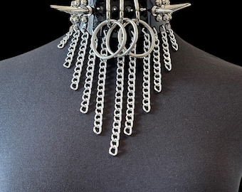 May include: A black glossy choker collar featuring silver-toned metal hardware, including two large side spikes, three central O-rings, and multiple dangling silver-toned chains of varying lengths that drape downwards from the front of the collar.
