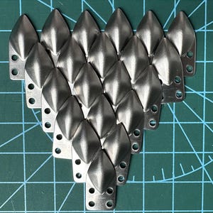 May include: Twenty-one silver metal scales, each with a pointed tip and a rectangular base with three holes, arranged in a heart shape. The scales are on a green grid surface. Each scale is approximately 2.5 cm long.