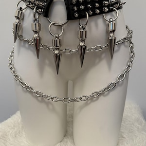 May include: Black leather belt with silver spikes and a chain detail. The belt features a large metal ring and dangling silver spikes. A silver chain hangs below the belt, adding a punk rock aesthetic.