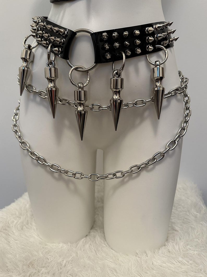 May include: Black leather belt with silver spikes and a chain detail. The belt features a large metal ring and dangling silver spikes. A silver chain hangs below the belt, adding a punk rock aesthetic.