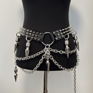 Könnte beinhalten: Ein schwarzer Gürtel mit zwei Reihen silberfarbener Metallspitzen, einem zentralen silbernen Ring und mehreren herabhängenden silbernen Ketten. Mehrere große, spitze silberfarbene Metallkegel hängen an den Ketten und bilden ein mehrlagiges Accessoire.