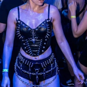 May include: A person in a crowded venue wearing a black leather corset with silver spikes and a matching belt with chain details. The outfit is accessorized with a chain belt and black shorts. The scene is illuminated with blue lighting.